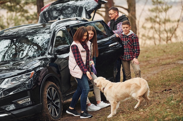 Фото Счастливая семья развлекается со своей собакой возле современного автомобиля на открытом воздухе в лесу