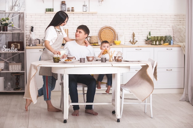 一緒に朝食を食べて幸せな家族。キッチンのテーブルで食べる若い家族。ママ、パパ、小さな赤ちゃんが食べています。