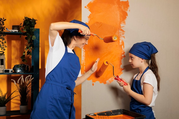 Happy family having fun painting at home, using orange paint color on roller paintbrush to redecorate apartment interior. Happy woman with little girl working on house renovation and decoration.