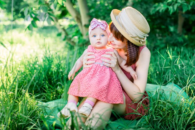 A happy family having a picnic in the green garden in a sunny spring day: a beautiful smiling mother sitting on green grass and her little laughing daughter on her legs