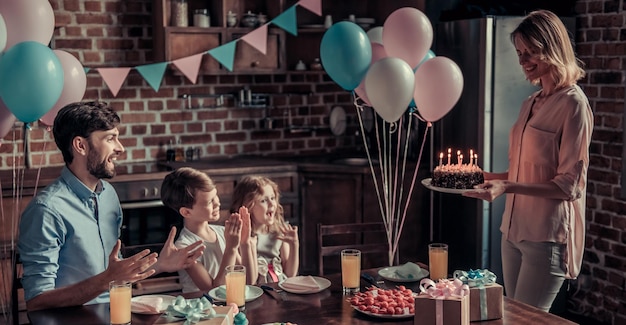 幸せな家族は誕生日のお祝いの間に装飾されたキッチンのテーブルに座っています、お母さんはバースデーケーキを持っています