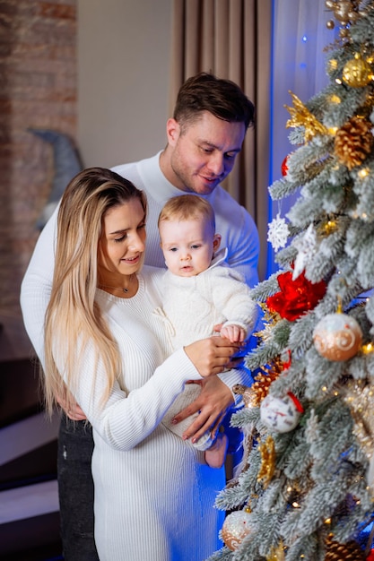 Happy family is standing near the Christmas tree and looking at the baby