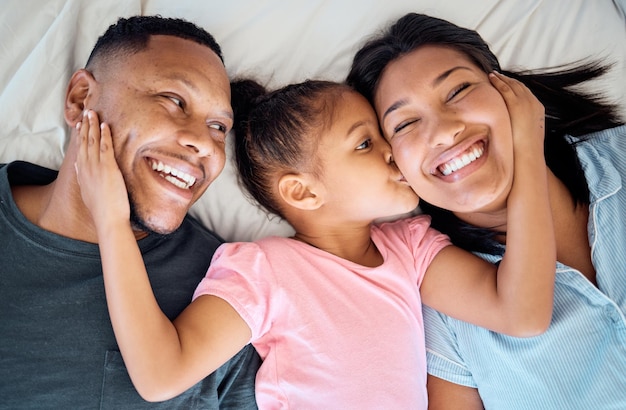 Happy family kid kiss and bed with a girl mother and father with a smile love and parent care Happiness hug and black family in a bedroom lying relax together in a morning with a child at home