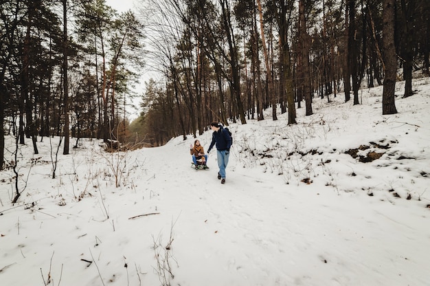 Famiglia felice: l'uomo trasporta la slitta di legno con la ragazza sulla slitta che cammina nella foresta all'aperto. passeggiate nel fine settimana di vacanza nel parco invernale.