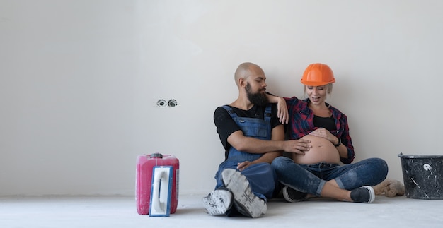 Happy family man and pregnant woman in hard hat husband and wife are resting while making repairs at home sitting by the wall and talking to their belly. White wall and room renovation concept.