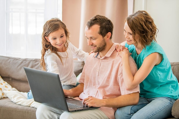 Happy family mom dad and daughter are sitting on couch and looking at laptop monitor together