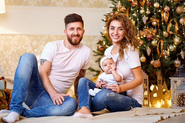 Happy family of mom dad and little baby girl sitting near Christmas tree on the floor