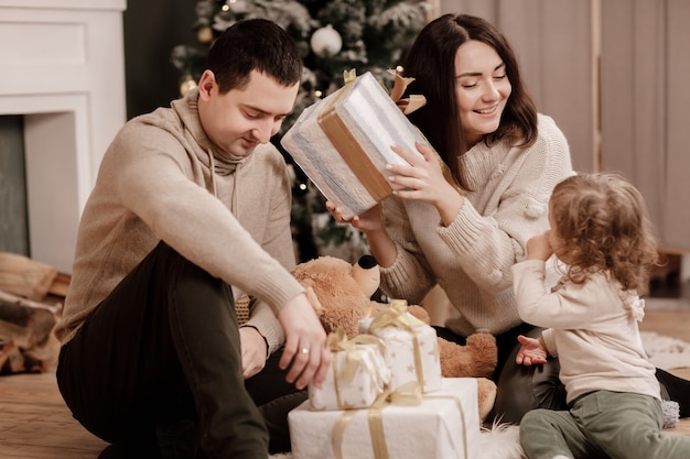 Happy family, mom, dad and little daughter with many presents boxes near Christmas tree and fire place at home.