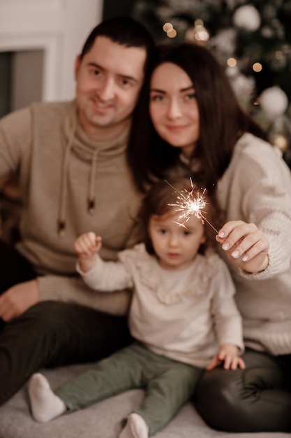 Happy family, mom, dad and little daughter with sparklers near christmas tree and fireplace at home.