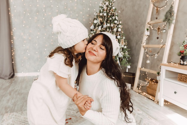 Happy family, mother and baby girl in white knitted hats sit near a Christmas tree with a Christmas present