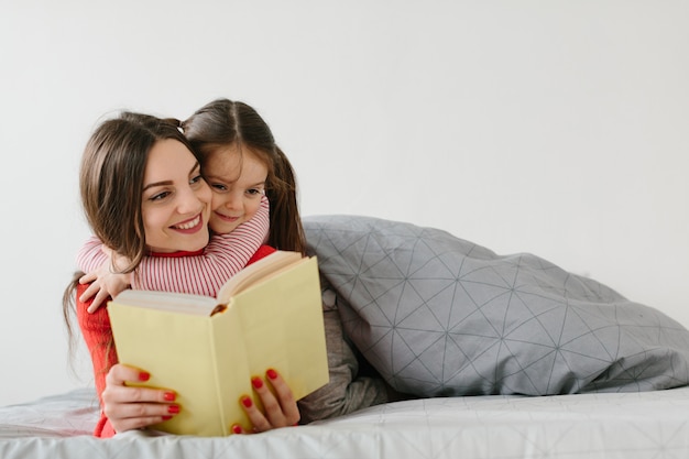 Happy family mother and child daughter reading holding book lying in bed