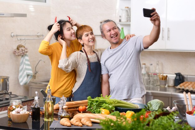 Happy family of mother father and daughter cooking in kitchen making healthy food together and using smart phone to take selfies