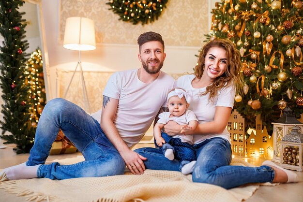 Happy family of mother father and little baby girl sitting near Christmas tree on the floor