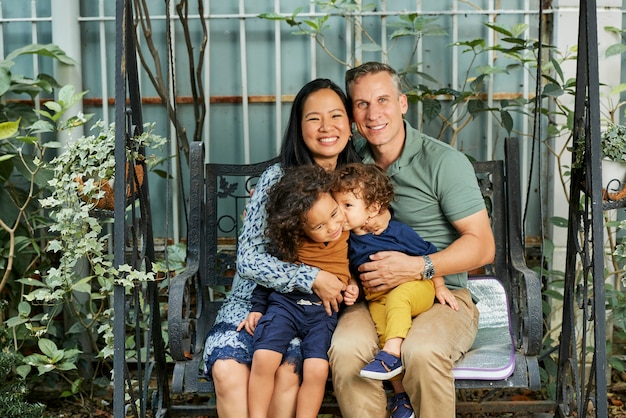 Happy family of mother, father and two little sons sitting on swing in backyard and smiling at camera