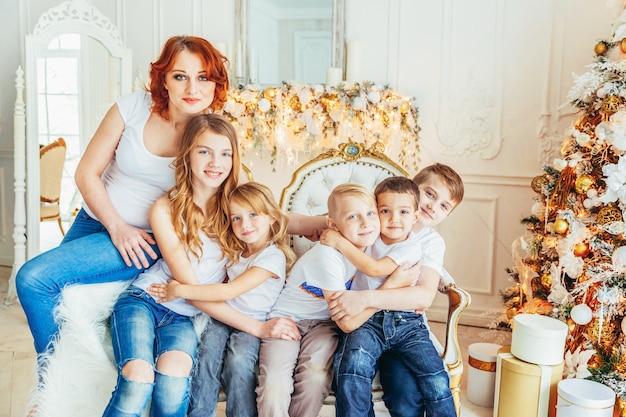 Happy family mother and five children relax playing near Christmas tree on Christmas eve at home. Mom, daughters, sons in light room with winter decoration. Christmas New Year time for celebration