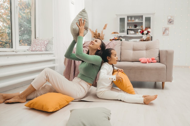 A happy family! Mother and her little daughter girl fight pillows on the floor near the sofa. Happy family games.