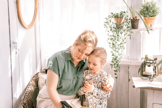 Happy family mother with cute little kid daughter girl have fun
together in suburb countryside home on sunny summer day candid mom
hugging and reads book to child exploring book with magnify
glass