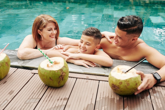 Счастливая семья из трех человек освежается в воде в солнечный летний день и пьет кокосовые коктейли