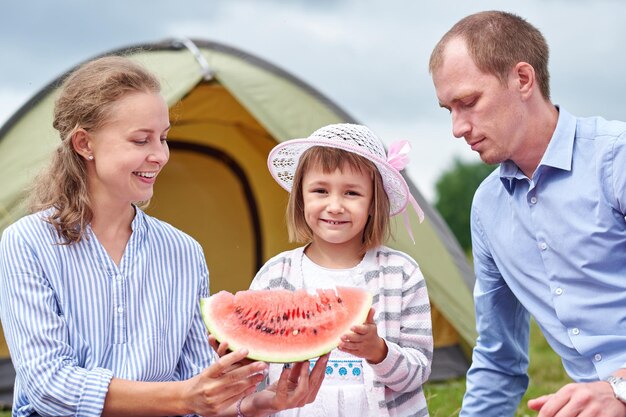 Happy family on picnic at camping Mother father and daughter eating watermelon near a tent in meadow or park