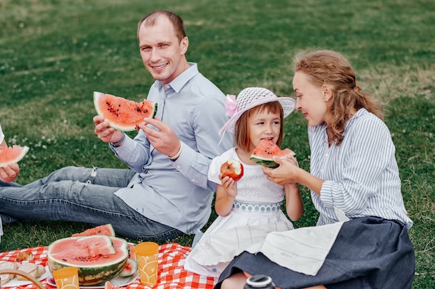 スイカを食べてピクニックで幸せな家族。牧草地や公園でのピクニックで母、父、子