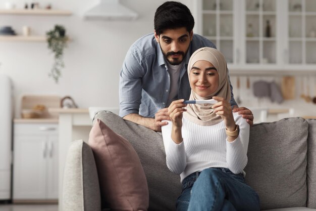 Happy family, planning baby, pregnancy and checking result at home. Glad cheerful young arab woman in hijab and man look at positive pregnancy test on sofa in living room interior, empty space