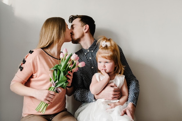 Ritratto di famiglia felice concetto amore di una vacanza in famiglia mamma con fiori papà abbracci figlia seduta in casa su uno sfondo chiaro emozioni di felicità festa della donna festa della mamma