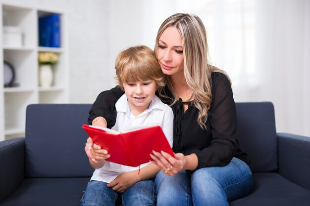 幸せな家族の肖像画-若い母親と彼女のかわいい幼い息子が家で一緒に本を読んでいます