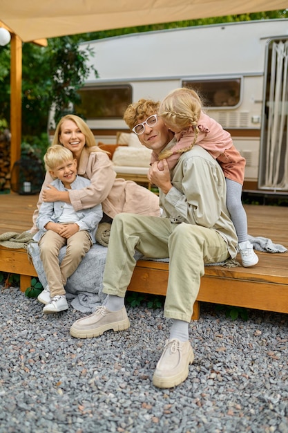 Happy family poses at the trailer, summer camping. Parents with children travel in camp car, nature and forest on background. Campsite adventure, travelling lifestyle