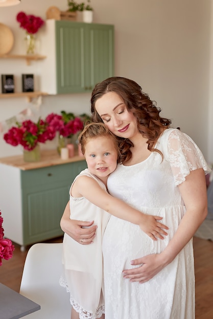 Happy family, pregnant woman with little daughter in kitchen with peonies flowers. Relationship between parents and children. Motherhood, pregnancy, happiness concept.