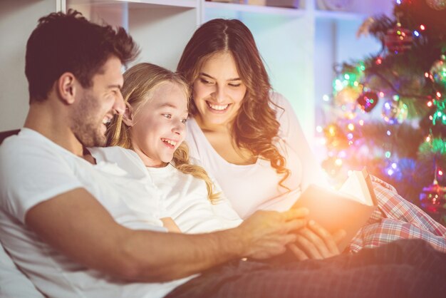 The happy family read the book in the bed near the christmas tree. evening time