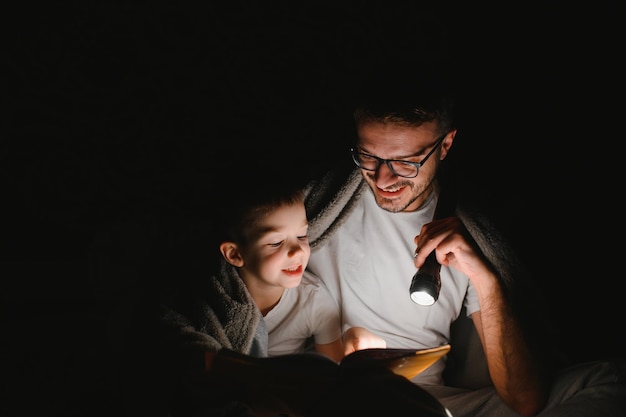 Happy family reading bedtime story under blanket in evening Father and son spend time together Father's Day