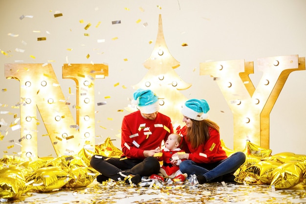 A happy family in red sweaters is sitting together on the floor. Christmas holiday atmosphere. A child in a santa costume. Family relationship concept