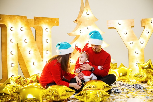 A happy family in red sweaters is sitting together on the floor. Christmas holiday atmosphere. A child in a santa costume. Family relationship concept