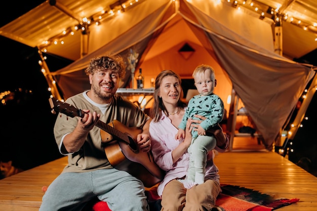 Happy family relaxing and spend time together in glamping on summer evening and playing guitar near cozy bonfire Luxury camping tent for outdoor recreation and recreation Lifestyle concept