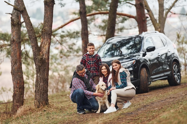 Happy family sitting and having fun with their dog near modern car outdoors in forest