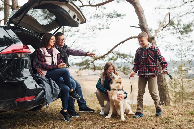 Happy family sitting and having fun with their dog near modern car outdoors in forest