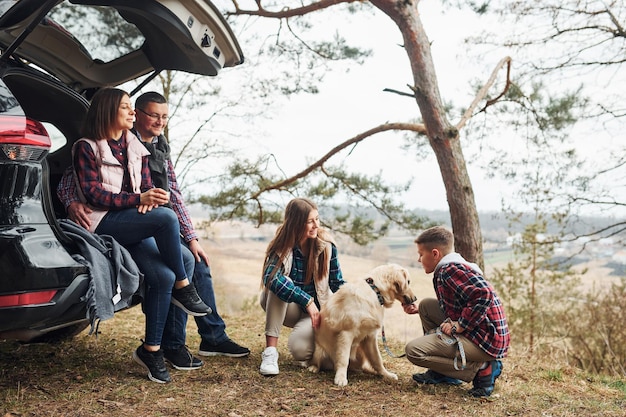 행복한 가족이 앉아서 야외에서 숲속의 현대적인 자동차 근처에서 개와 즐거운 시간을 보낸다