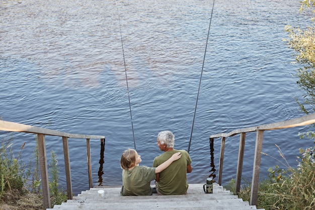 Happy family spending time together in open air near river or lake, son hugging his father with love while sitting on wooden stairs leading to water.