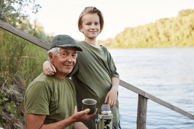 Happy family spending time together outdoor. Father and son sitting on wooden placing near water, drinking hot beverage, boy hugging his dad.