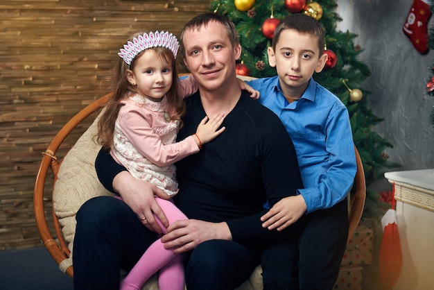 Happy family spends time together on winter vacation at home by the fireplace near the Christmas tree with gifts. Cute little girl and boy with his father in the chair at the Christmas tree.