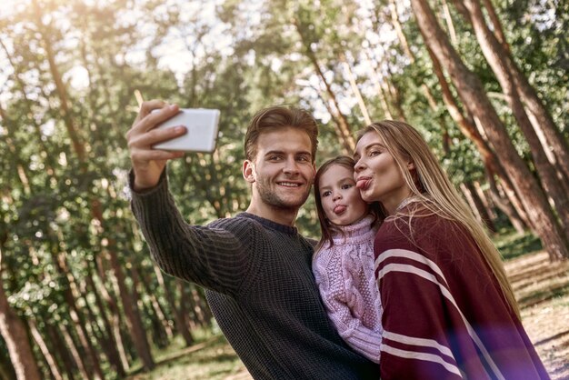 Счастливая семья, делающая селфи с помощью смартфона в лесу. Отец держит смартфон с камерой