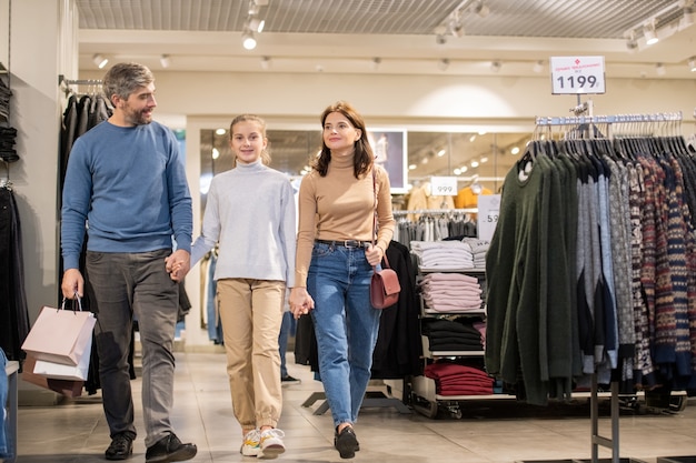 Happy family of three in casualwear holding by hands while moving along clothing department during seasonal sale period