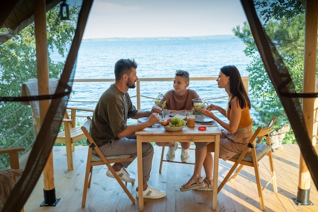 Happy family of three having dinner by wooden table on terrace