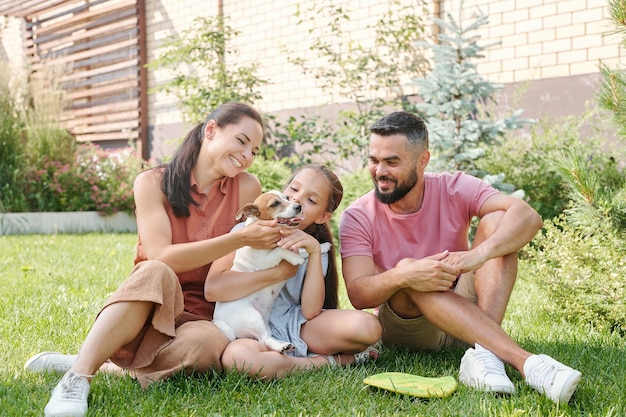 Happy family of three spending time together with their little dog in backyard sitting on lawn