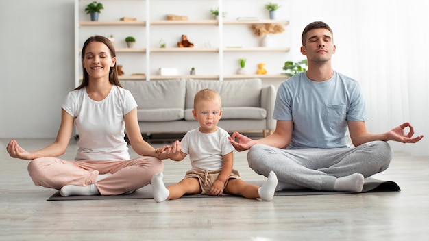 Happy Family Of Three With Little Infant Baby Meditating Together At Home, Smiling Millennial Parents And Their Adorable Toddler Son Sitting In Lotus Position, Enjoying Practicing Yoga, Copy Space