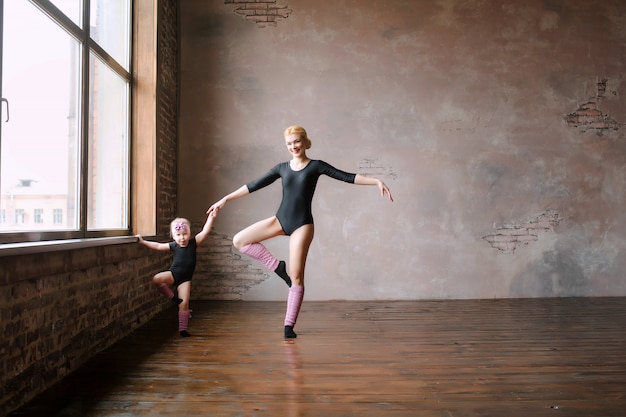 счастливая семья, две балерины, мать и дочь в черных гимнастических купальниках у розовой стены