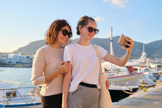 Happy family on vacation, mom and teenage daughter taking selfie on smartphone, sea bay with yachts summer sunset background