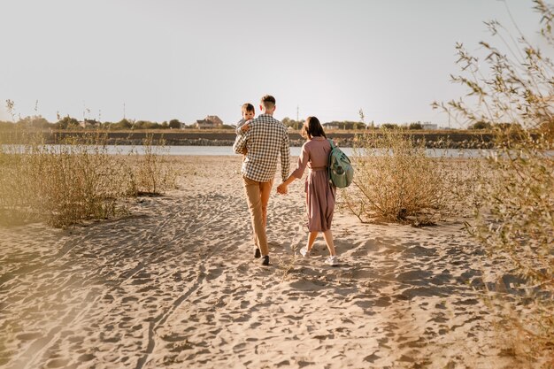 川の砂浜を歩いて幸せな家族。父、母は赤ん坊の息子を手に持って一緒に行きます。背面図。ファミリータイズのコンセプト。