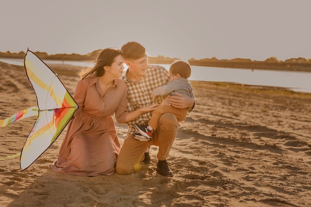 Happy family walking on sandy beach of river. Father, mother holding baby son on hands and playing with kite.