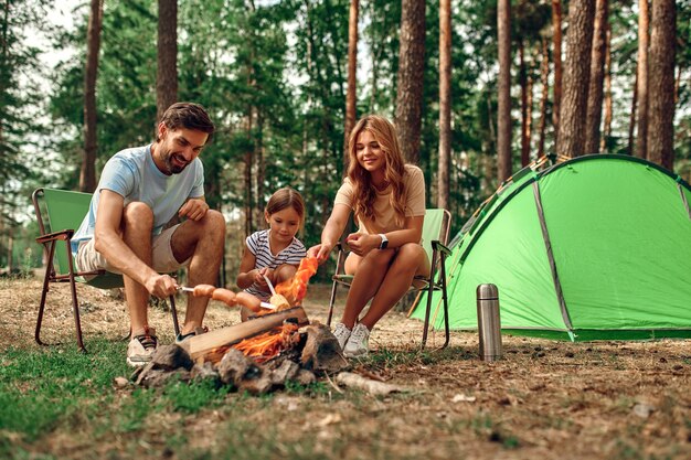 Happy family with a child on a picnic sit by the fire near the\
tent and grill a barbecue in a pine forest. camping, recreation,\
hiking.